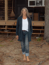 A woman stands barefoot in front of a rustic log cabin porch, wearing a dark Navy open-front Blondie Classic Briton Cardigan over a white t-shirt, cuffed blue jeans, and a brown belt.