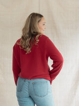 A woman with blonde hair is shown from the back wearing a Poppy red long-sleeve crewneck Blondie Classic Camp Crew Sweater and light blue jeans, standing against a neutral beige fabric backdrop.