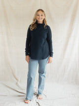 A woman stands barefoot in front of a draped cream fabric backdrop, wearing a dark navy long-sleeve Blondie Classic Dusk Sweater and light blue jeans.