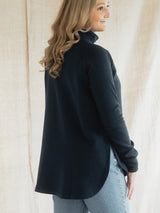 A woman wearing a navy long-sleeve Blondie Classic Dusk Sweater and light wash jeans, photographed from a side-back angle against a neutral beige fabric backdrop.