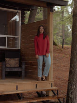 A woman stands on a rustic wooden cabin porch wearing a Poppy red Blondie Classic Dusk Sweater, light blue jeans, and sandals, with trees and a wooded outdoor setting in the background.