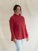 A woman wearing a Blondie Dusk Sweater top in a Poppy red waffle knit fabric and light blue jeans, standing against a neutral beige fabric backdrop.