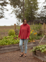 A woman wearing a Blondie Dusk Sweater top in a Poppy red waffle knit fabric, light blue jeans, and black sandals stands in a gravel path of a garden with raised wooden beds and green plants.