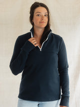 A woman wearing a navy blue quarter-zip long-sleeve Blondie Mountaineer Sweater with a white zipper trim, paired with light blue jeans, standing against a neutral off-white fabric backdrop.