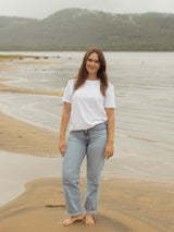 A woman stands barefoot on a sandy beach wearing a plain white short-sleeve crewneck Blondie Apparel Prairie t-shirt and light-wash blue jeans, with calm water and a forested hill in the background.