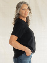 A woman with gray curly hair wearing a black short-sleeve Blondie Apparel Prairie t-shirt and blue jeans, standing against a light beige fabric backdrop.