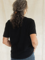 A woman viewed from behind wearing a black short-sleeve Blondie Apparel Prairie t-shirt and blue jeans, with her hair in a low bun secured with an orange hair tie, against a neutral cream-colored fabric backdrop.