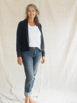 A woman stands barefoot against a draped off-white fabric backdrop, wearing a dark navy Blondie Spurce Cardigan over a white top with blue jeans and a brown belt.