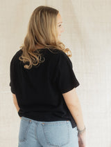 A woman with blonde hair is shown from the back wearing a black short-sleeve Blondie tide tee in slub cotton fabric and light blue jeans, standing against a neutral textured beige backdrop.
