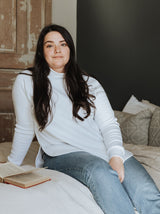 woman sitting on a bed wearing a classic dusk sweater in white