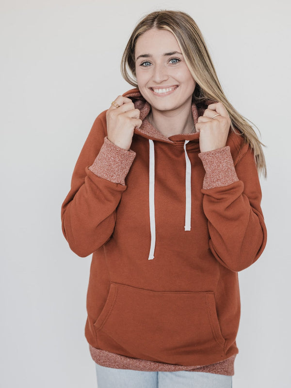 Blonde woman smiling, holding hood while wearing the Blondie Classic Forest Hoodie in Gingerbread Red with heather cuffs, hem and lining