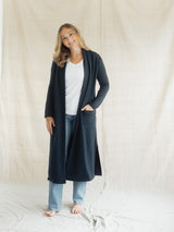A woman modeling a long, dark navy blue open-front Blondie Apparel Aspen Cardigan over a white t-shirt and light blue jeans, standing against a draped beige fabric backdrop.