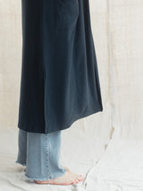 A person wearing a long dark navy draped Blondie Apparel Aspen Cardigan over light blue frayed-hem jeans, standing barefoot against a draped off-white fabric backdrop.