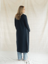 A woman with long wavy light brown hair is shown from the back wearing a long, dark navy blue Blondie Apparel Aspen Cardigan over light-wash wide-leg jeans. She is standing in front of a draped, off-white fabric backdrop.