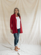 A woman stands barefoot in front of a draped beige fabric backdrop, wearing a bright Poppy red Blondie Briton Cardigan over a white top and blue jeans.