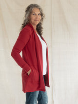 A woman with gray curly hair models a Poppy red open-front Blondie Briton Cardigan with side pockets over a white top and blue jeans, standing against a draped beige fabric backdrop.