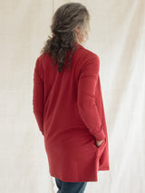 A woman with long, wavy gray-brown hair is shown from behind wearing a long-sleeve Poppy red Blondie Briton Cardigan with side pockets, paired with blue jeans, against a draped off-white fabric backdrop.