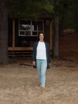 A woman walking barefoot on sandy ground in front of a rustic wooden cabin surrounded by pine trees, wearing a white t-shirt, light blue jeans, and a dark navy Blondie Briton Cardigan.