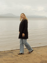 A woman stands on a sandy shore next to a calm body of water under an overcast sky, wearing a black long-sleeve Blondie Classic Briton Cardigan, light blue jeans, and black sandals.