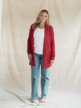 A woman stands barefoot in front of a draped beige fabric backdrop, wearing a bright Poppy red open-front Blondie Classic Briton Cardigan over a white top and light blue jeans.