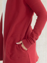 A close-up of a person wearing a long-sleeve Poppy red Blondie Classic Briton Cardigan, with one hand tucked into a front patch pocket. The fabric is smooth and the shot is cropped tightly against a light neutral background.