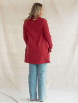 A woman is shown from the back wearing a Poppy red long-sleeve Blondie Classic Briton Cardigan and light blue jeans, standing against a draped beige fabric backdrop.