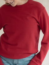 A close-up of a person wearing a solid Poppy red crewneck Blondie Classic Camp Crew Sweater with light blue jeans, cropped from the chin down against a neutral light background.