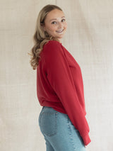 A woman wearing a Poppy red Blondie Classic Camp Crew Sweater and light blue jeans, standing against a draped beige fabric backdrop.