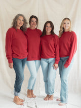 Four women standing together and wearing matching Poppy red Blondie Classic Camp Crew Sweater with light-wash blue jeans, posed against a draped off-white fabric backdrop.