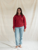 A woman stands barefoot in front of a draped off-white fabric backdrop, wearing a Poppy red long-sleeve Blondie Classic Coastal Sweater with a button placket and light blue jeans.