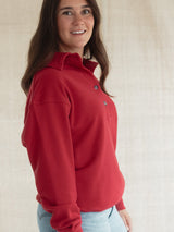 A woman wearing a Poppy red long-sleeve Blondie Classic Coastal Sweater with light blue jeans, standing against a neutral beige textured background.