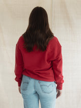 A person viewed from behind wearing a Poppy red long-sleeve Blondie Classic Coastal Sweater and light blue jeans, standing against a plain beige fabric backdrop.