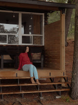 A woman sits barefoot on the wooden porch steps of a rustic cabin in a forest setting, wearing a Poppy red Blondie Classic Fauna Sweater and light blue jeans.