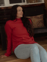 A woman with long dark hair sits on a wooden floor wearing a bright Poppy red Blondie Classic Fauna Sweater and light blue jeans in a rustic indoor setting.