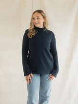 A woman modeling a dark navy blue Blondie Classic Fauna Sweater with light blue jeans, standing against a neutral beige fabric backdrop.