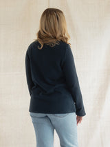 A woman viewed from behind wearing a dark navy long-sleeve Blondie Classic Fauna Sweater and light blue jeans, standing against a neutral beige fabric backdrop.