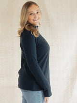 A woman with blonde hair wearing a dark navy Blondie Classic Fauna Sweater and light blue jeans, standing against a neutral beige fabric backdrop.