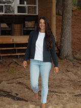 A woman walking barefoot on a dirt path in front of a cabin, wearing a dark navy blue Blondie Classic Spruce Cardigan over a white t-shirt and light-wash blue jeans.