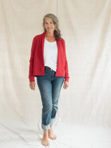 A woman stands barefoot against a draped cream fabric backdrop, wearing a bright Poppy red open-front Blondie Classic Spruce Cardigan a white top with blue jeans.