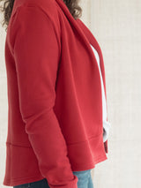 A close-up side view of a person wearing a Poppy red cropped Blondie Classic Spruce Cardigan or cardigan with a white side panel, paired with blue jeans against a light neutral wall.
