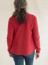 A woman viewed from behind wearing a Poppy red long-sleeve Blondie Classic Spruce Cardigan and blue jeans, standing against a light beige fabric backdrop.