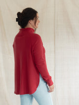 A woman wearing a Blondie Dusk Sweater top in a Poppy red waffle knit fabric and light blue jeans, shown from a side-back angle against a neutral cream fabric backdrop.