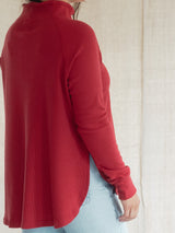 A person wearing a Blondie Dusk Sweater top in a Poppy red waffle knit fabric with light blue jeans, shown from a side-back angle against a neutral beige fabric backdrop.