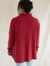 A woman viewed from behind wearing a Blondie Dusk Sweater top in a Poppy red waffle knit fabric and light blue jeans, standing against a neutral beige fabric backdrop.