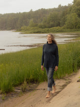 A woman walking barefoot on a dirt path next to a grassy marsh with water and a forest in the background, wearing a Blondie Dusk Sweater top in a navy blue waffle knit fabric and gray rolled-up jeans.