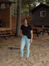 A woman standing outdoors in a wooded setting, wearing a black Blondie Janis t-shirt  and light-wash blue jeans.