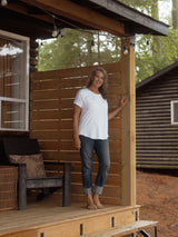 A woman stands barefoot on a wooden cabin porch wearing a white Blondie Janis t-shirt and dark gray rolled-up jeans, with a wooden fence and trees in the background.