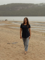 A woman walking on a sandy beach towards the camera, wearing a dark navy blue Blondie Janis t-shirt and light gray jeans, with a lake and forested hills in the background.