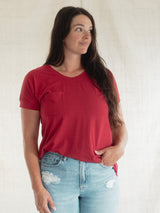 A woman wearing a Poppy red Blondie Janis t-shirt and light blue distressed jeans, standing against a light beige textured backdrop.