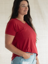 A woman wearing a Poppy red Blondie Janis t-shirt and light blue distressed jeans, standing against a light beige textured backdrop.